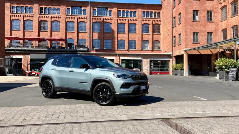 Silver Jeep Compass with black wheels parked in a sunny red brick industrial courtyard under clear skies.
