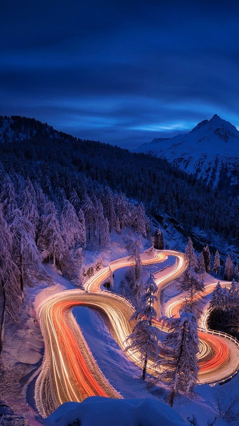 Warm light trails curve along a snow-covered mountain road under a dark blue twilight sky with evergreen trees.