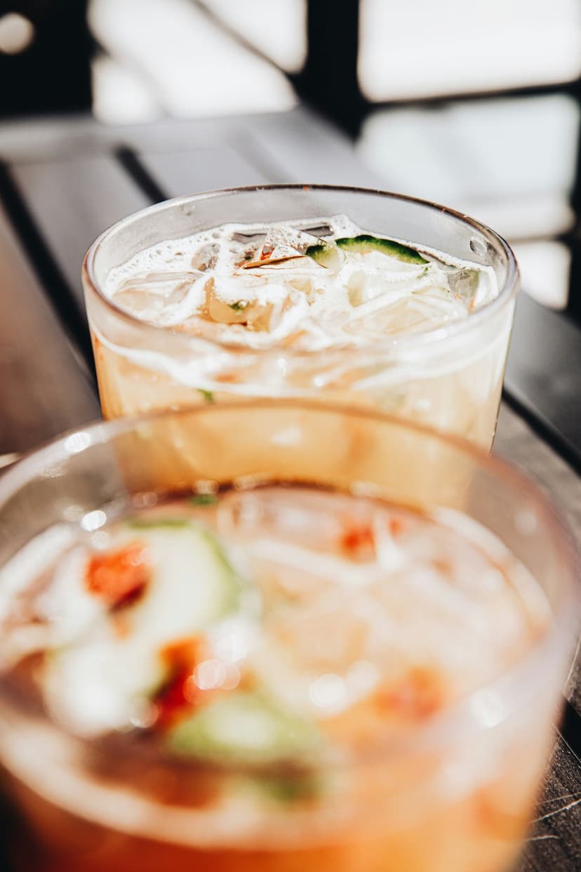 Two glasses of amber cocktails with ice cubes and cucumber slices set against a soft, blurred background.