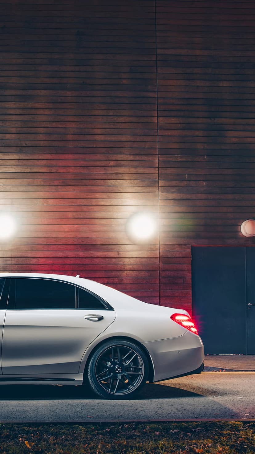 White luxury sedan parked against a wooden wall at night with glowing amber sconces and red taillights visible.