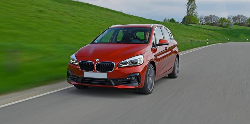Red BMW 2 Series Active Tourer driving on a winding road past green hills with motion blur effect.