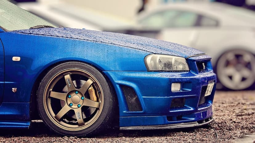 Blue Nissan Skyline GT-R R34 front fender with bronze wheels and raindrops on the paint in a wet setting.