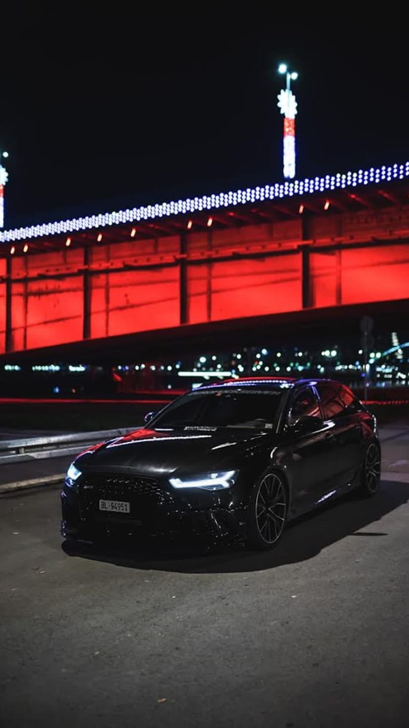 Black Audi RS6 Avant with glowing headlights parked on a city street under a red illuminated bridge at night.
