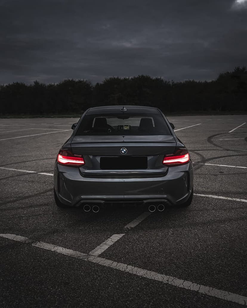 BMW M2 Coupe rear view with illuminated red taillights on a dark asphalt lot under a cloudy twilight sky.