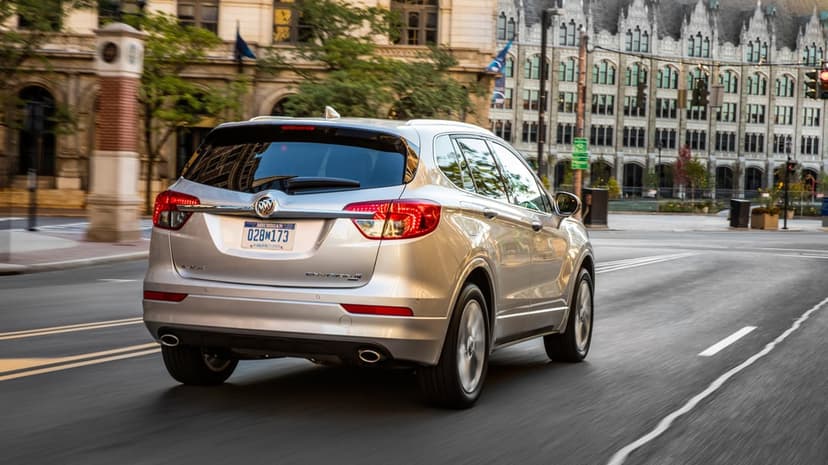 Silver Buick Envision SUV driving on a city street past historic buildings with motion-blurred road and wheels.