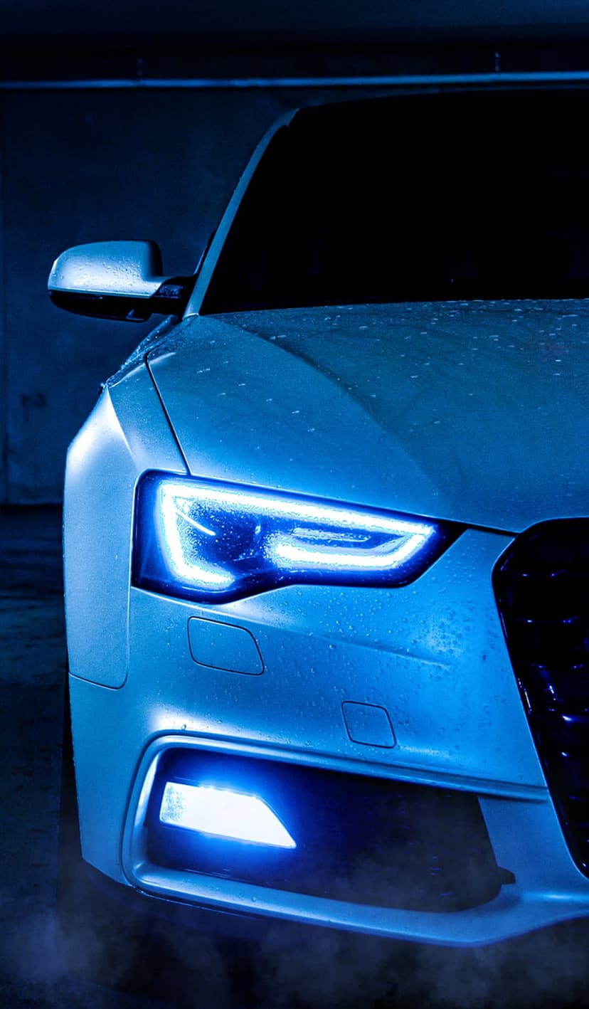 Blue LED headlights on a white Audi with water droplets reflecting light in a dark parking garage environment.