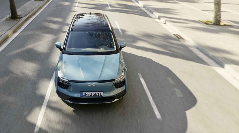 Top view of a light blue electric SUV driving on an urban street with long tree shadows in bright daylight.