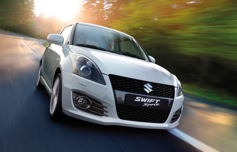 White Suzuki Swift Sport car driving fast on a winding road through a green forest at golden hour sunset.
