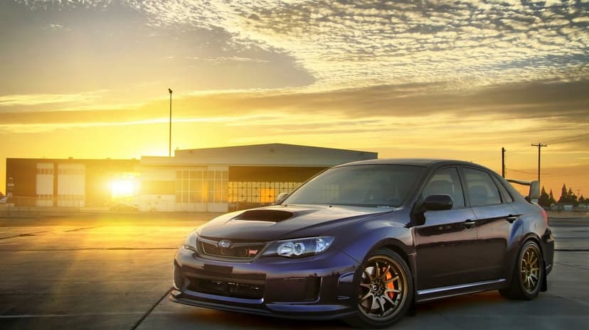 Purple Subaru WRX STI with bronze wheels parked on an airfield tarmac at sunset with a bright sun flare.