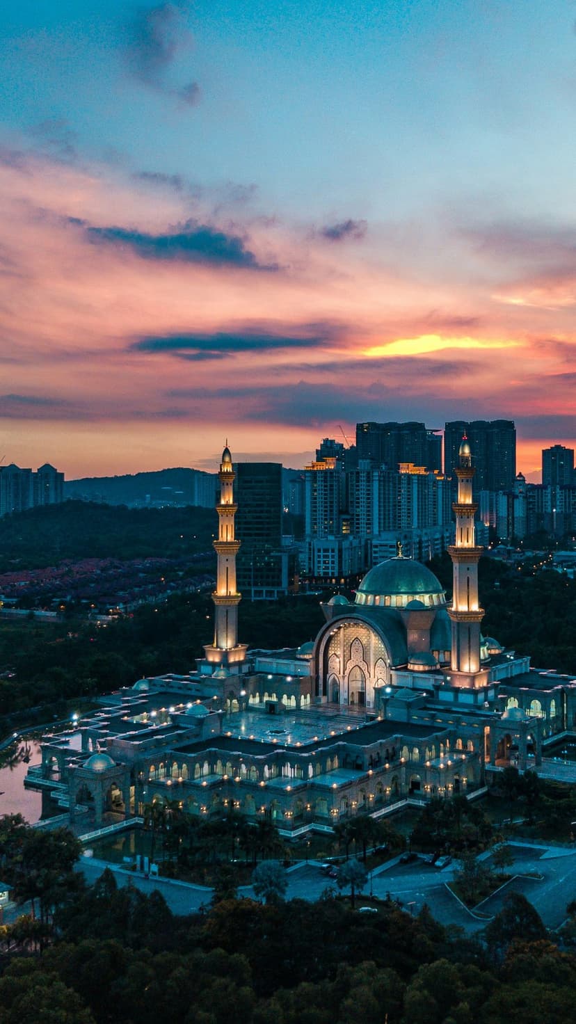 Grand mosque minarets reflect in water beside a metropolitan skyline during a vibrant orange and purple sunset.