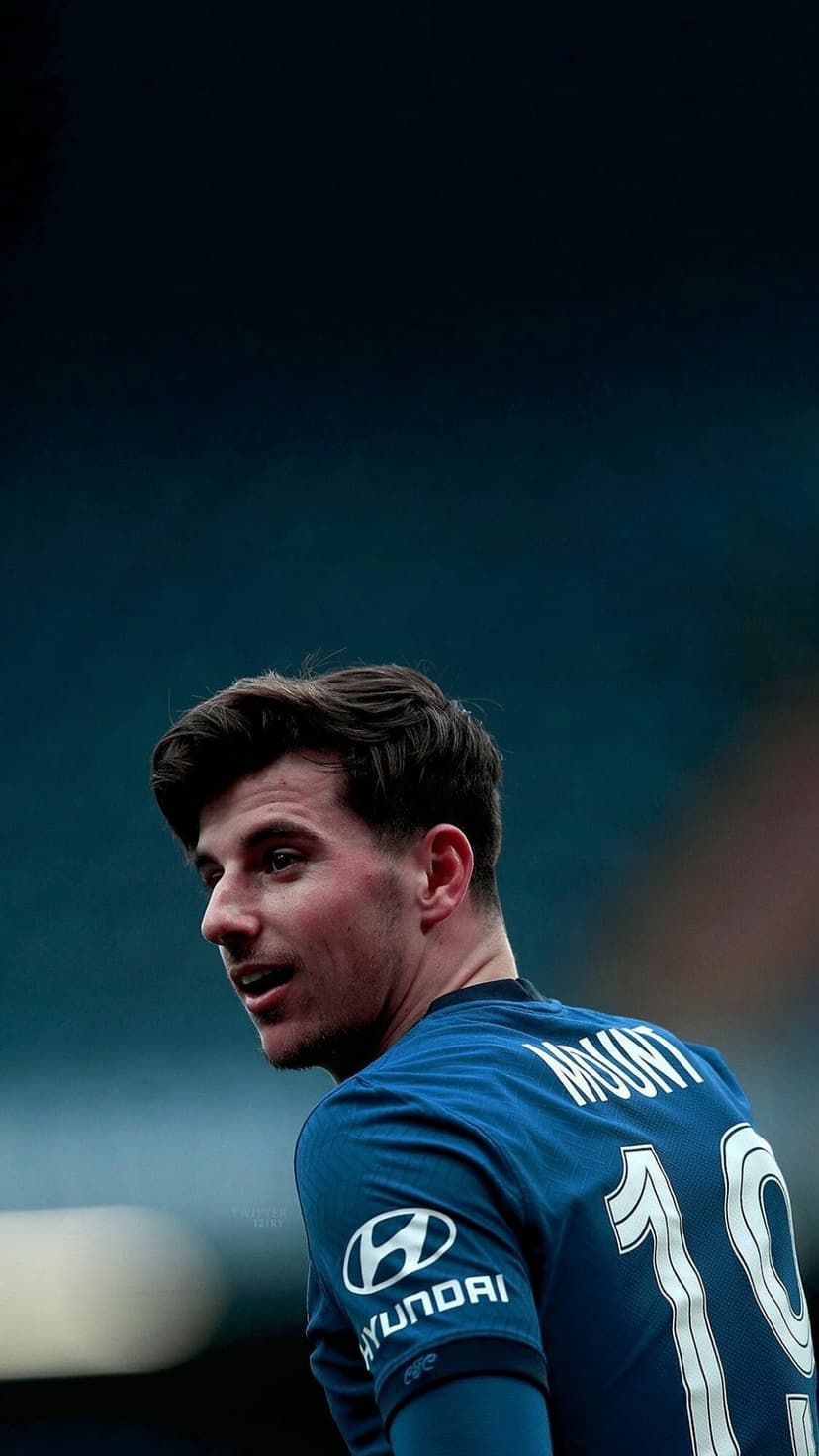 Mason Mount in a blue Chelsea jersey looks aside with a smile against a blurry dark blue stadium background.