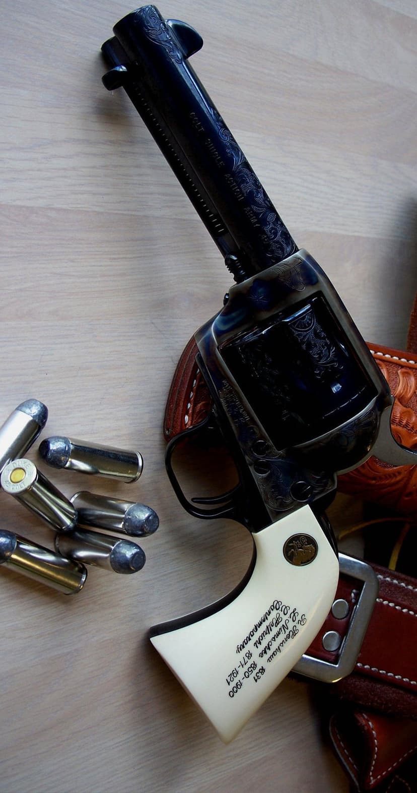 Engraved Colt Single Action Army revolver with ivory grips on a wooden surface next to leather gear and bullets.