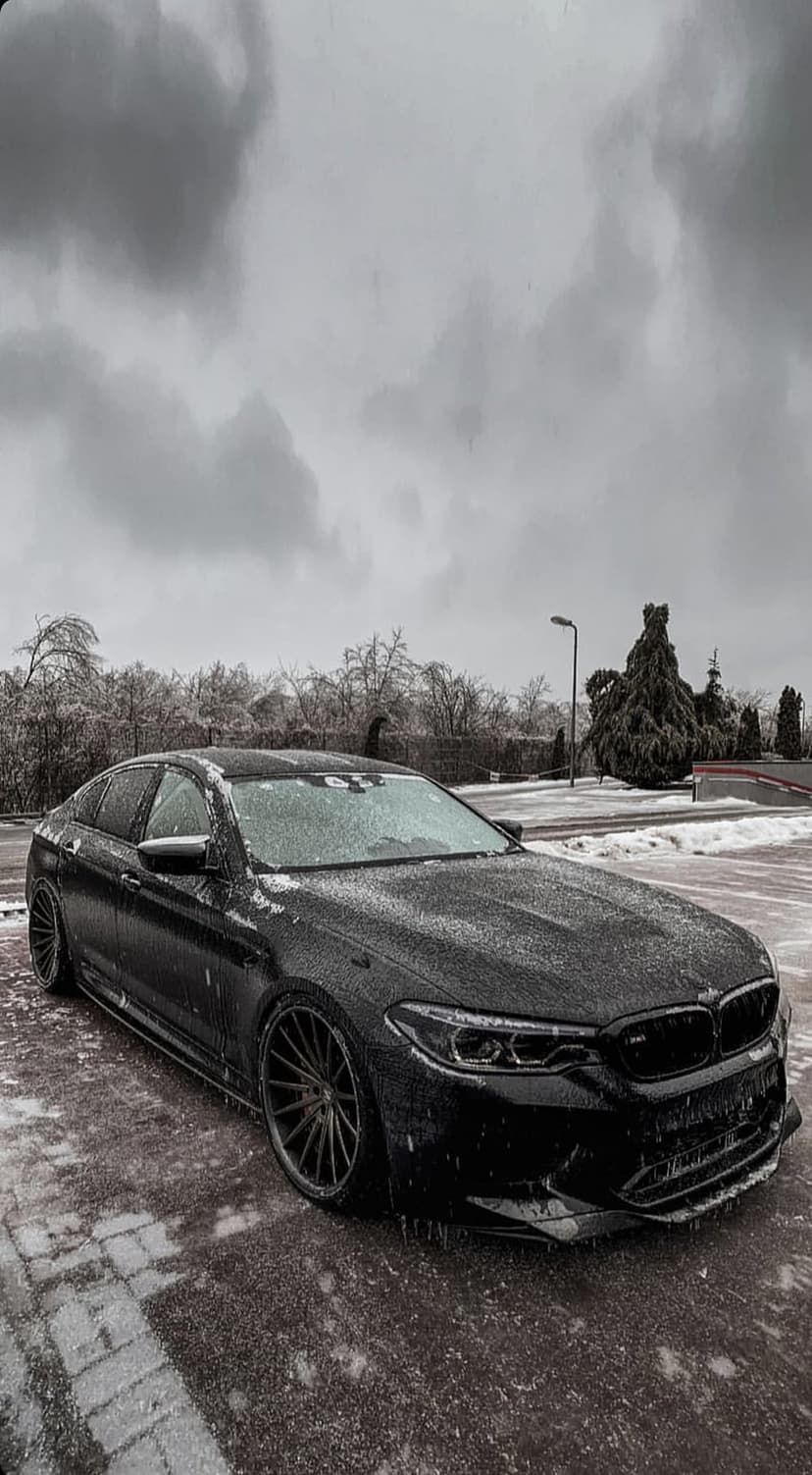 Black BMW M5 sedan parked on ice-slicked cobblestones surrounded by snow-draped trees under a cloudy winter sky.