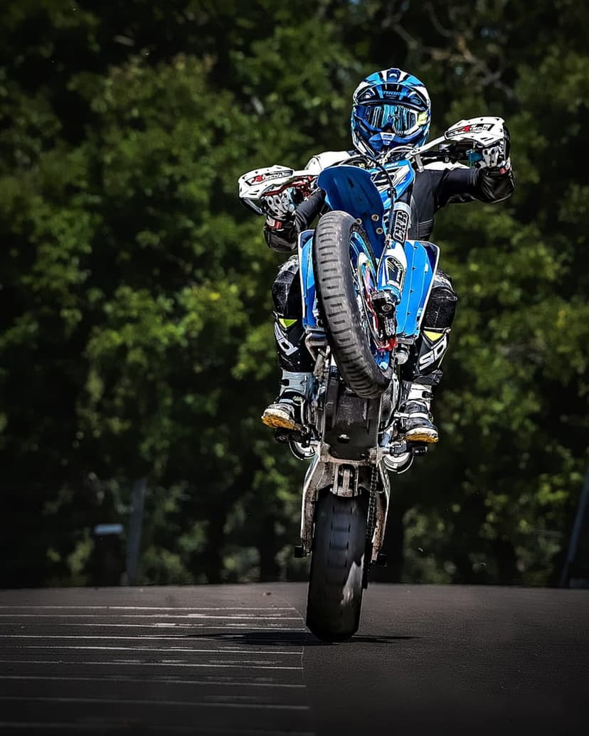 Motorcyclist in blue and white gear performs a vertical wheelie on a dirt bike along a tree-lined asphalt road.