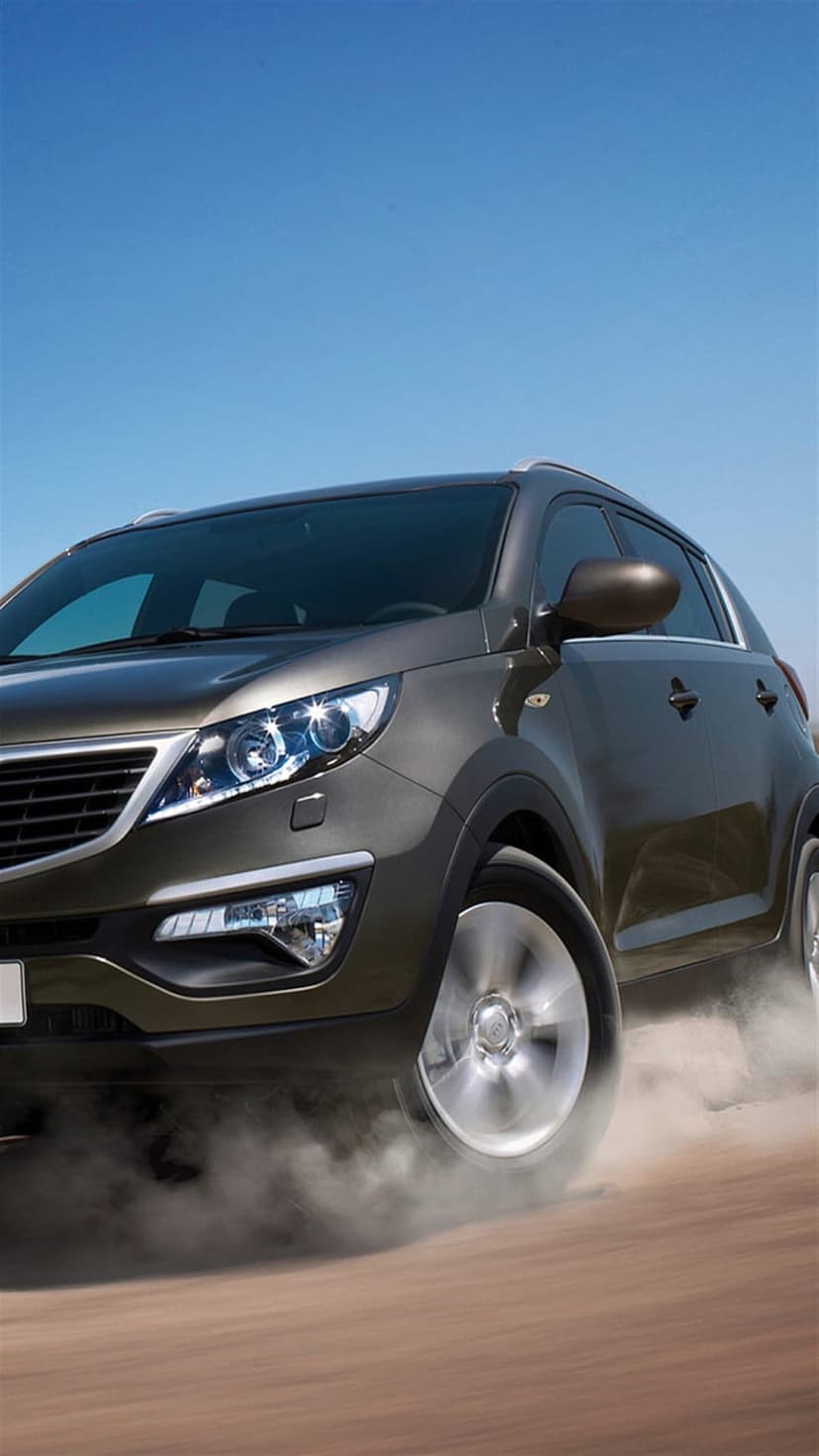Dark SUV speeds along a dirt trail with thick dust clouds rising against a clear blue sky in bright daylight.