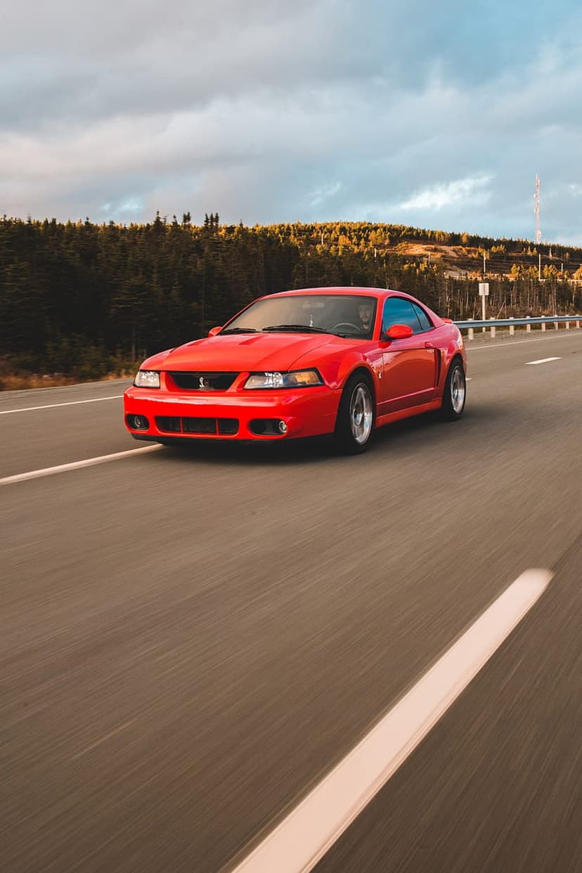 Red Ford Mustang GT driving on an asphalt highway surrounded by green trees under a cloudy sky at a low angle.