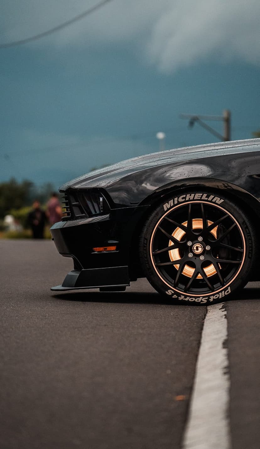Black Ford Mustang front wheel showing white Michelin tire lettering and gold brake calipers on asphalt.