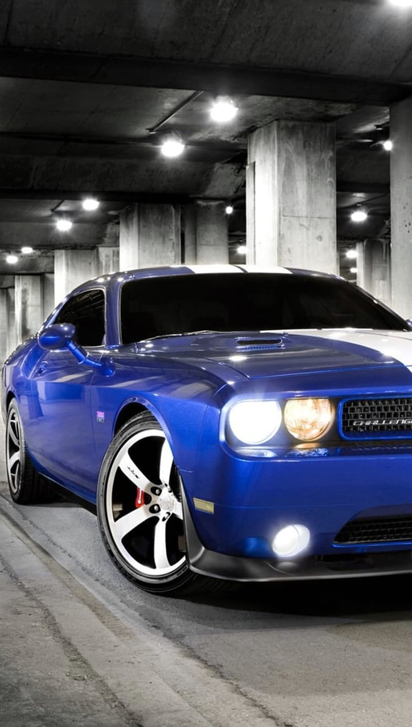 Blue Dodge Challenger with white stripes and glowing headlights parked in a concrete industrial garage.