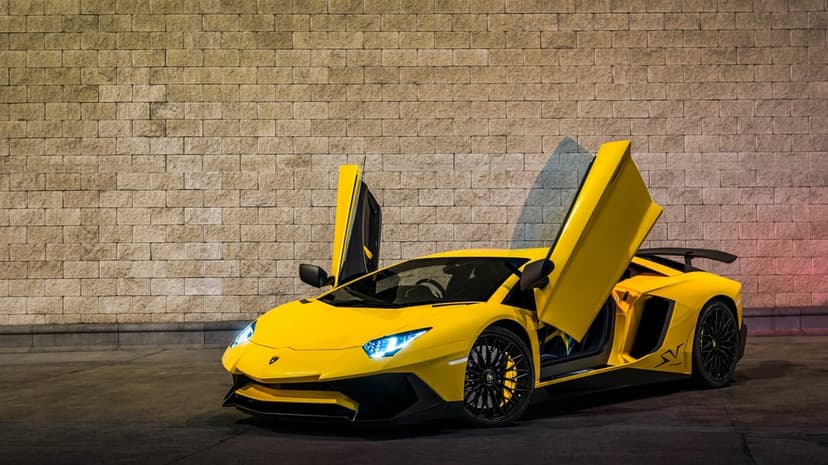Yellow Lamborghini Aventador SV with scissor doors open against a tan stone wall under evening lights.