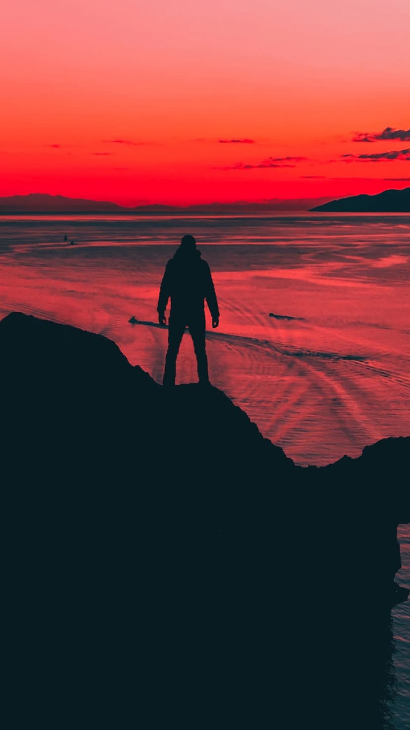 Lone silhouette stands on a rugged cliff overlooking a fiery red sunset reflecting across vast ocean waters.