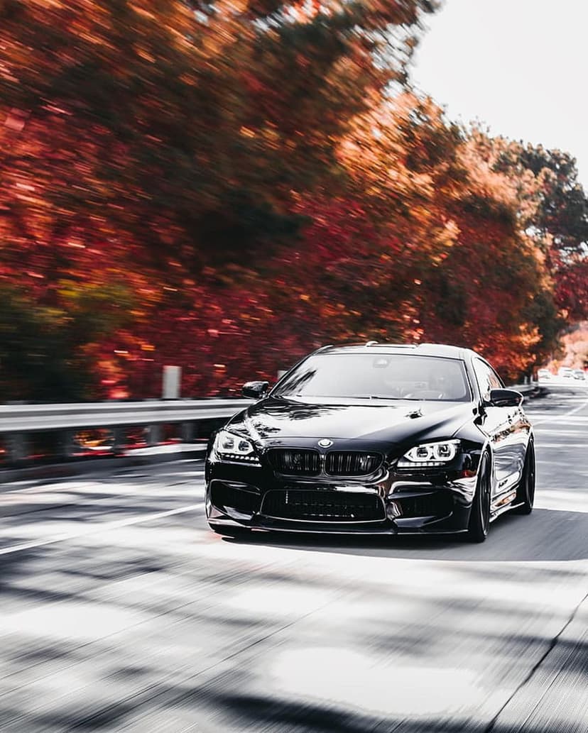 BMW M6 coupe in black driving fast past bright orange autumn trees with cinematic motion blur on a paved road.