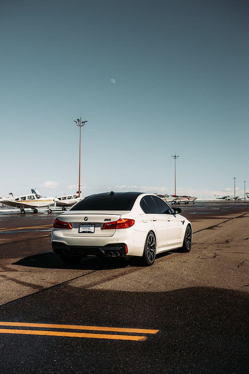 White BMW M5 sedan on an asphalt airport tarmac with propeller planes and a blue sky in the background.