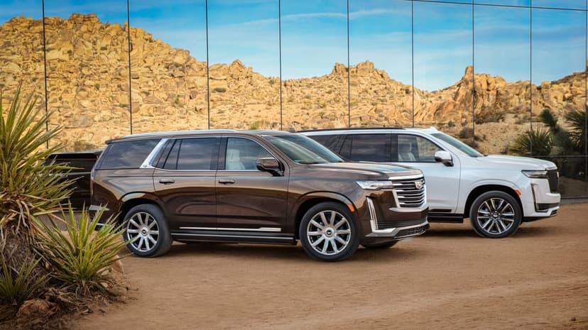Dark bronze and white Cadillac Escalades parked on sand before a reflective glass wall and desert hills.