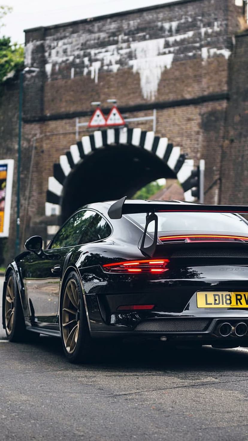 Black Porsche 911 GT3 with gold rims and a rear wing driving through a brick archway tunnel at night.