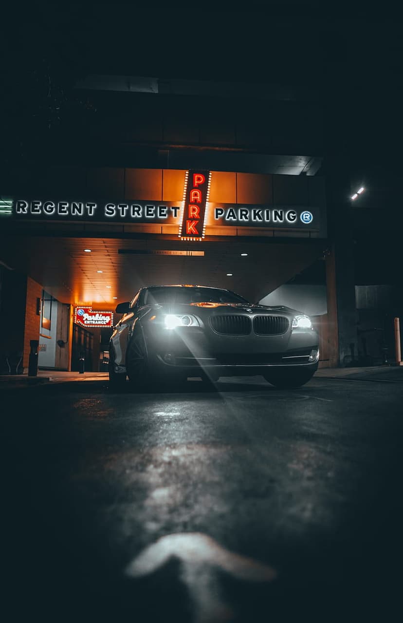 Modern car parked at a neon-lit parking entrance with bright headlights and reflections on wet city pavement.
