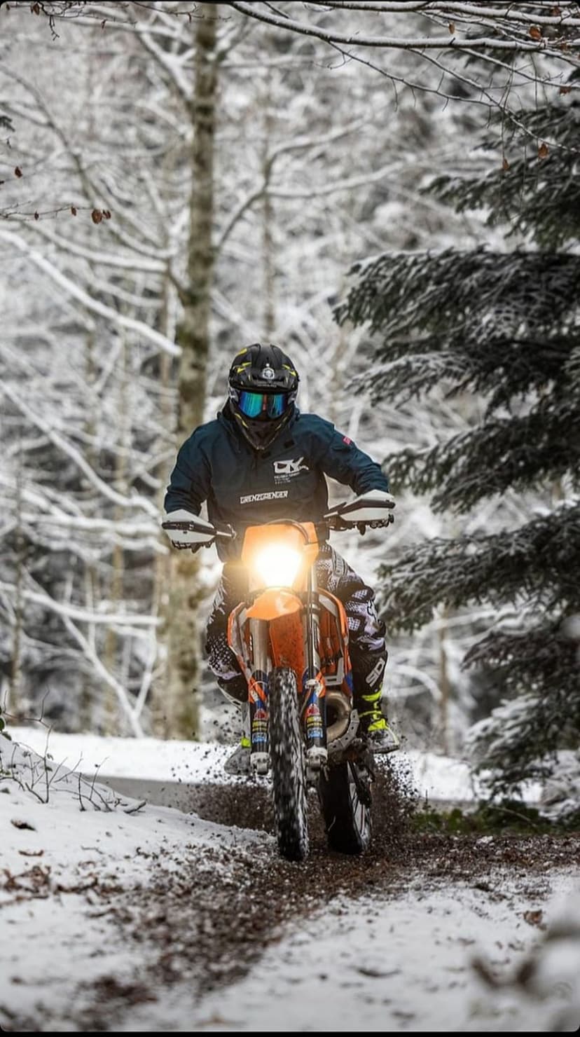 Rider on an orange dirt bike spraying snow and mud while racing through a dense, white winter forest trail.