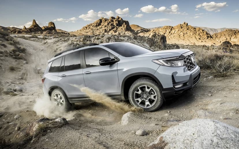 Silver SUV kicks up a dust cloud while driving on a rocky desert trail with mountains under a bright cloudy sky.