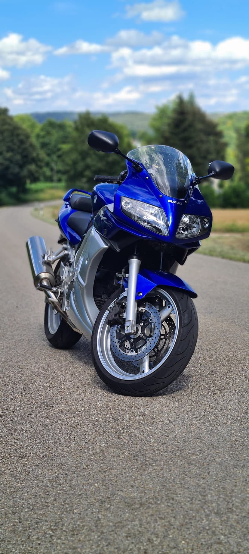 Blue Suzuki sportbike with dual headlights parked on an asphalt road overlooking green hills under a blue sky.