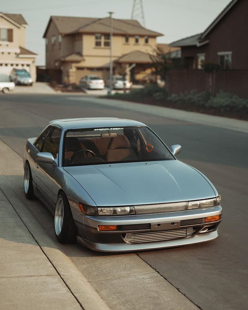 Silver Nissan Silvia S13 with aftermarket wheels parked on a suburban street under warm afternoon sunlight.
