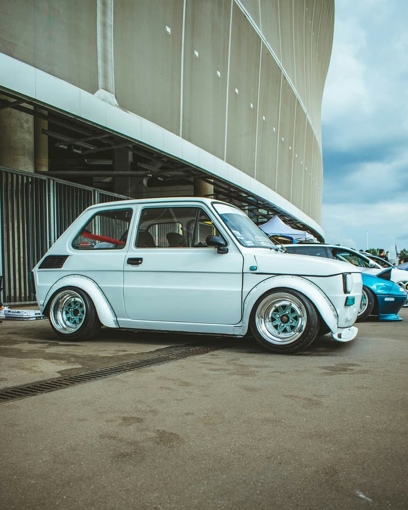 White Fiat 126p with wide body fenders and blue wheels parked on wet pavement near a modern curved building.