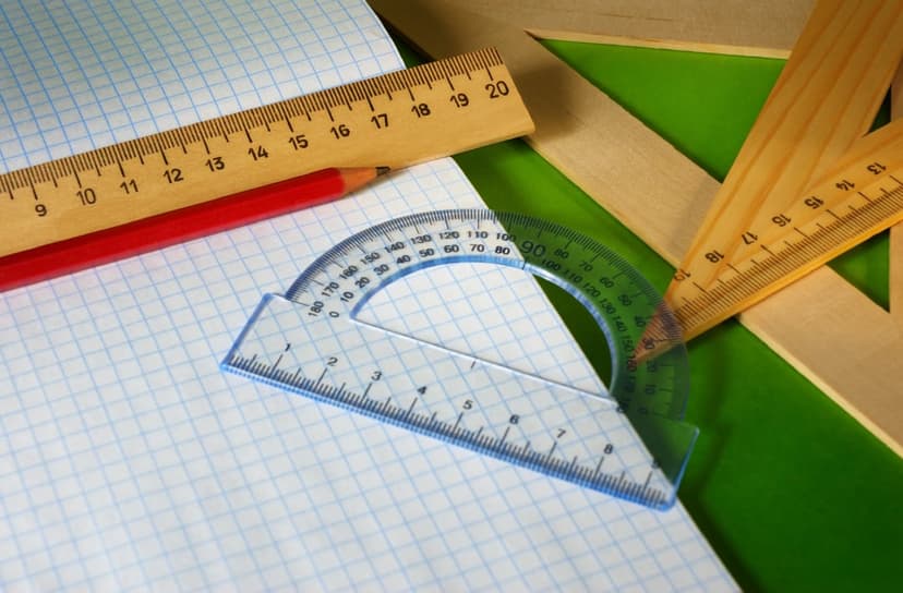 Wooden rulers, a protractor, and a red pencil lie on graph paper over a vibrant green background.