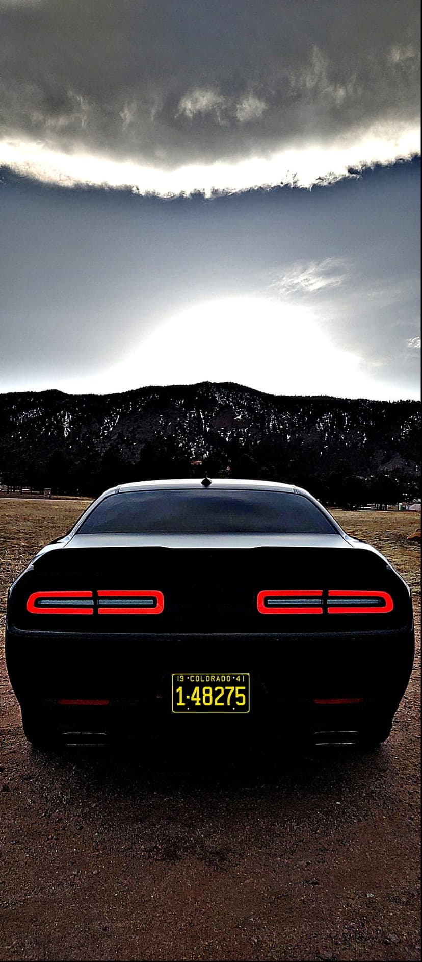 Black Dodge Challenger rear with glowing red LED tail lights and a yellow license plate under a cloudy mountain sky.