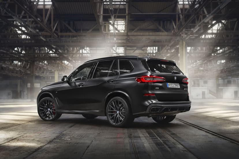 Matte black BMW X5 SUV parked in a sunlit industrial warehouse with dramatic overhead lighting and steel beams.