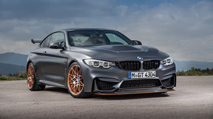 BMW M4 GTS matte grey sport coupe with orange rims and carbon fiber details on an asphalt road under a cloudy sky.