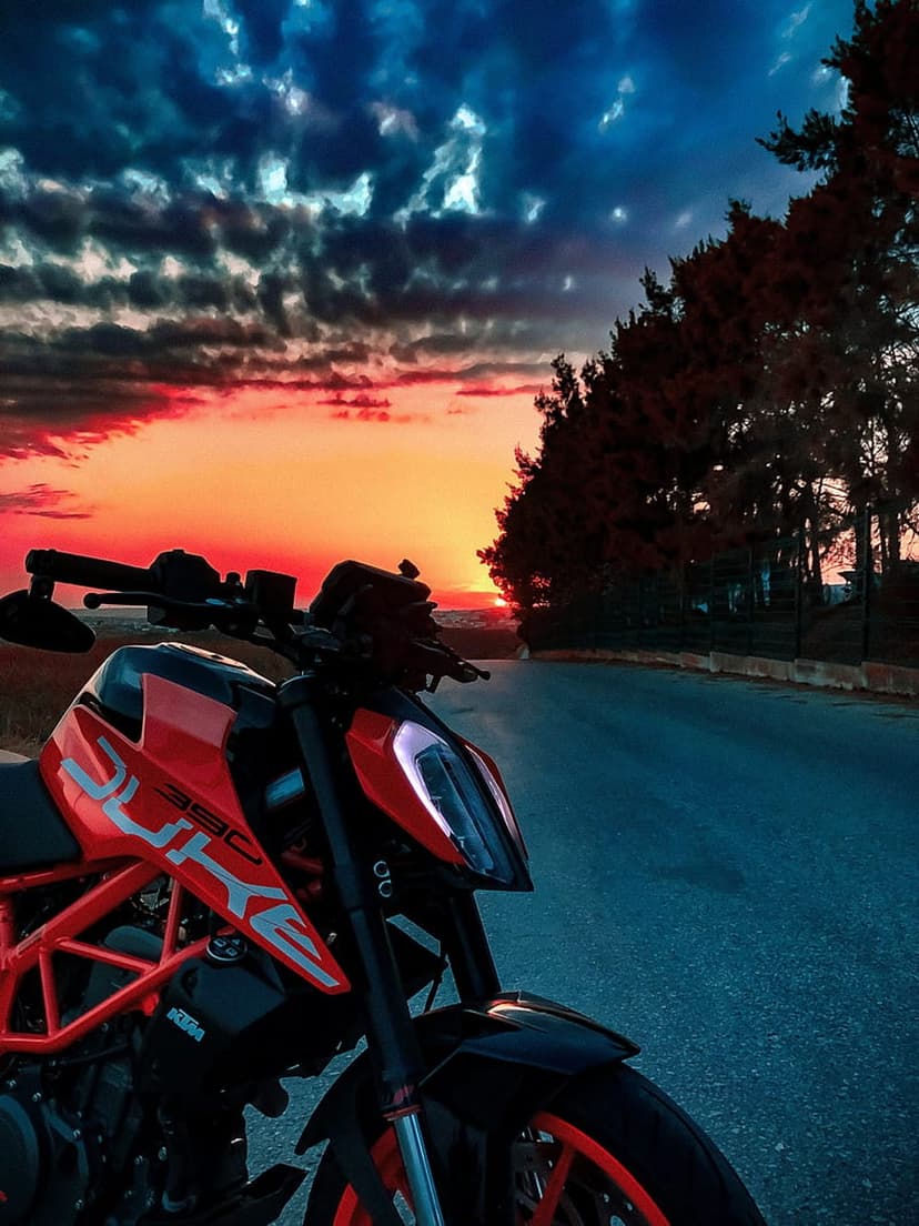 KTM Duke 390 motorcycle parked on an asphalt road under a dramatic orange sunset sky with lit headlights.
