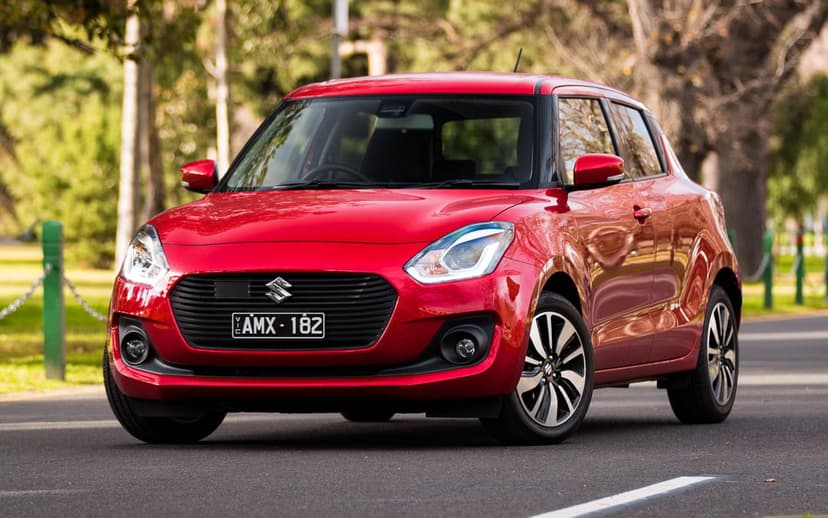 Red Suzuki Swift hatchback parked on a quiet asphalt road with blurred green park trees in the background.