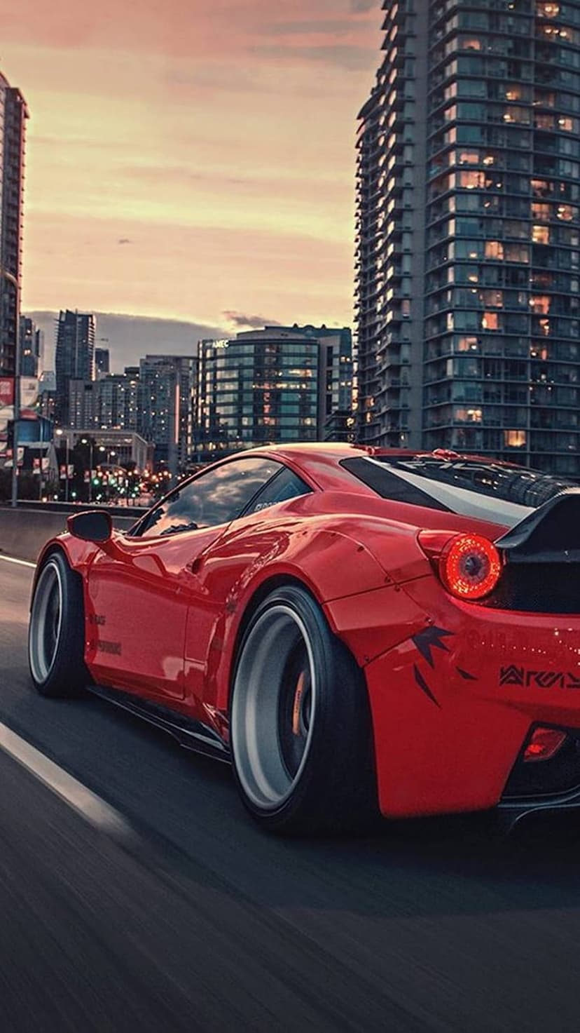 Red Ferrari sports car driving fast on an urban highway during a vibrant orange sunset with tall buildings.