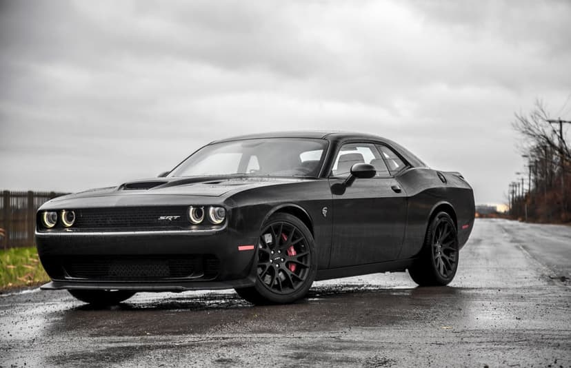 Black Dodge Challenger SRT Hellcat parked on a wet asphalt road under a cloudy sky with red brake calipers.