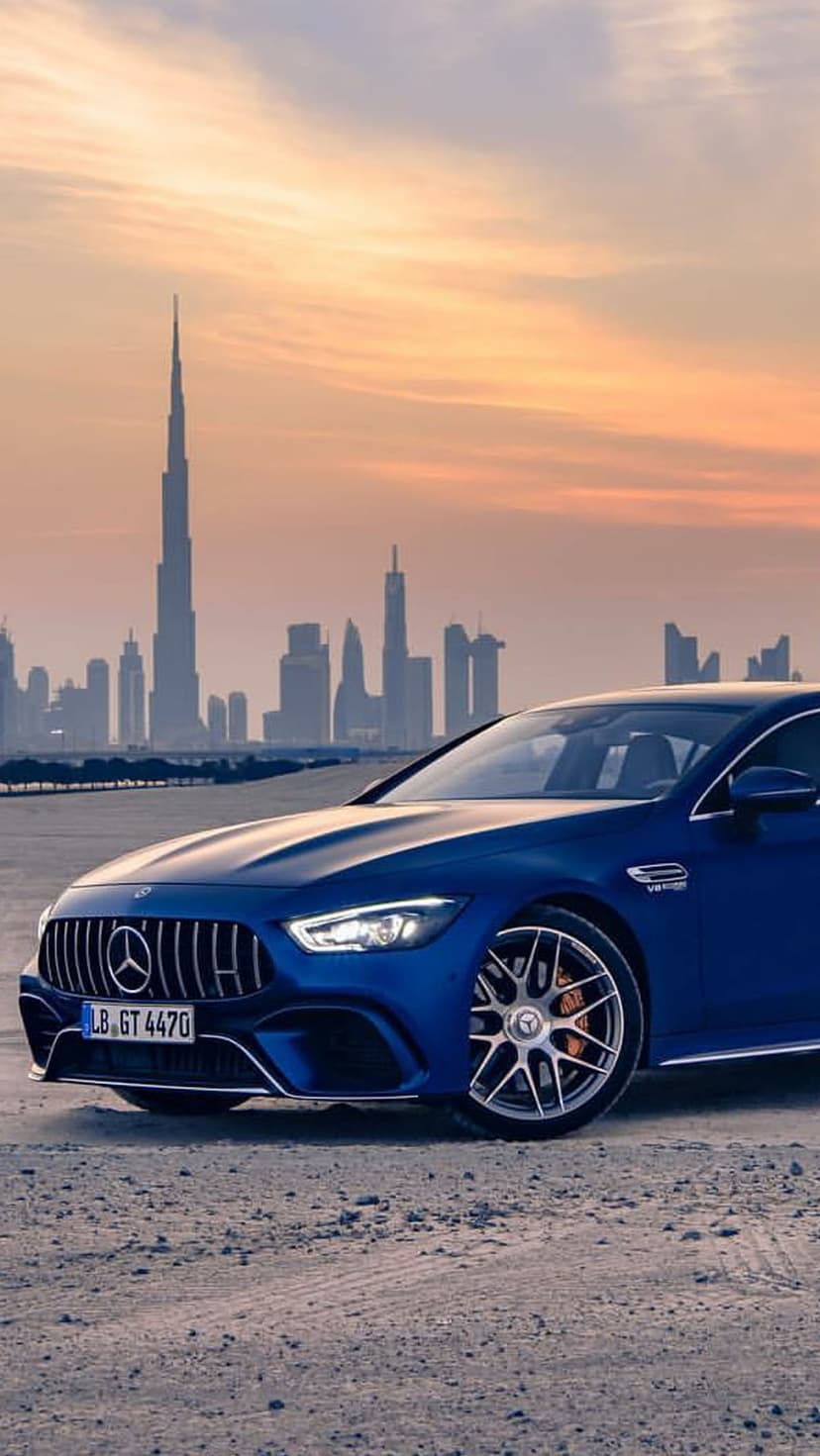 Blue Mercedes-AMG GT 63 S parked on sand with the Burj Khalifa and Dubai skyline during a vivid orange sunset.