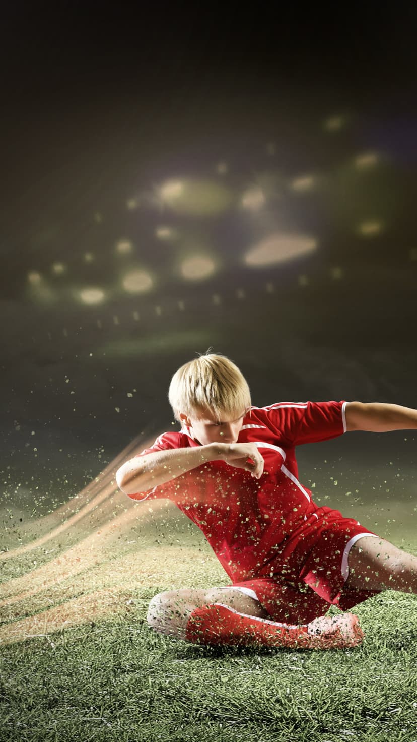 Young soccer player in a red jersey kicks a football with grass spraying under bright stadium floodlights.