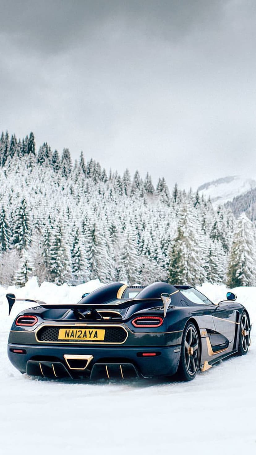 Dark blue Koenigsegg hypercar with gold accents parked on a snowy road surrounded by frosted evergreen trees.