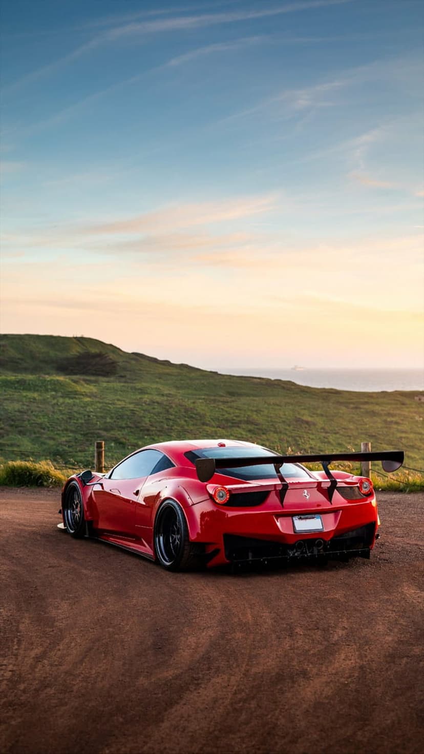 Red Ferrari 458 Italia with a large spoiler parked on a dirt path overlooking the ocean at sunset.