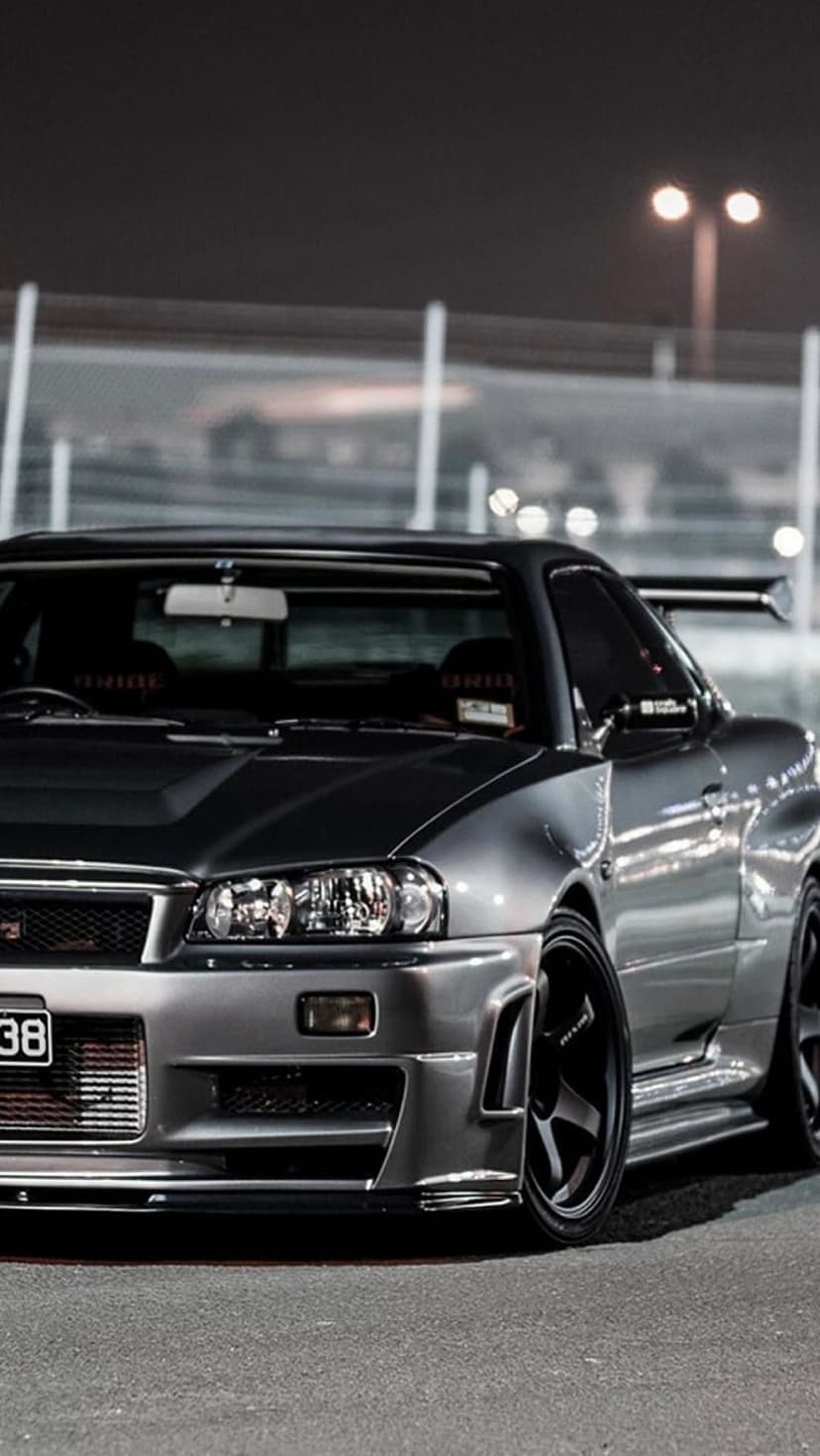 Silver Nissan Skyline R34 GT-R with Bride seats and a large rear wing parked under evening streetlights.