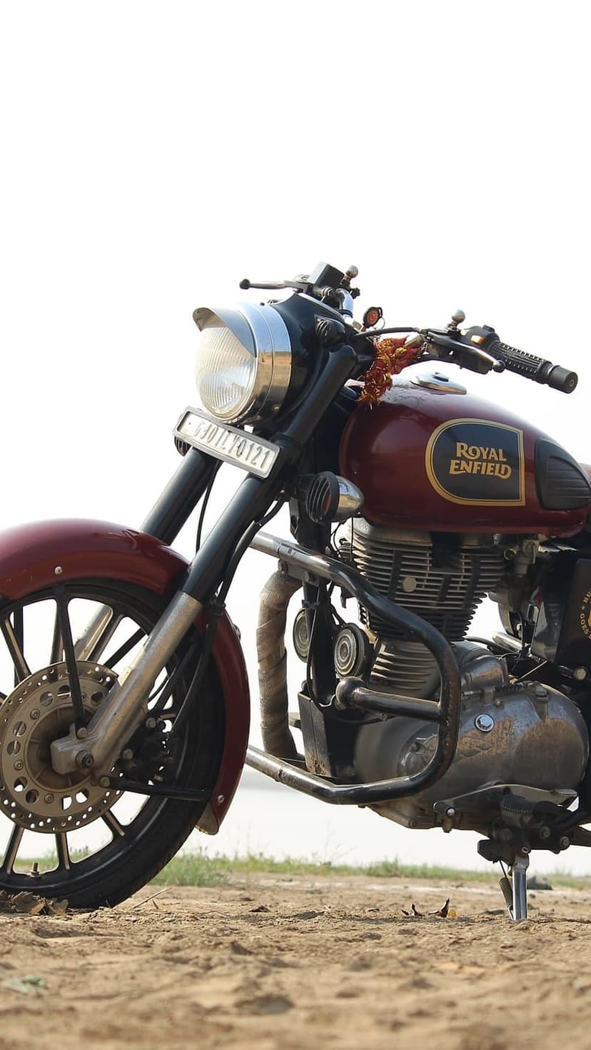 Maroon Royal Enfield motorcycle with a chrome engine and logo parked on sandy terrain near a blurred coastline.