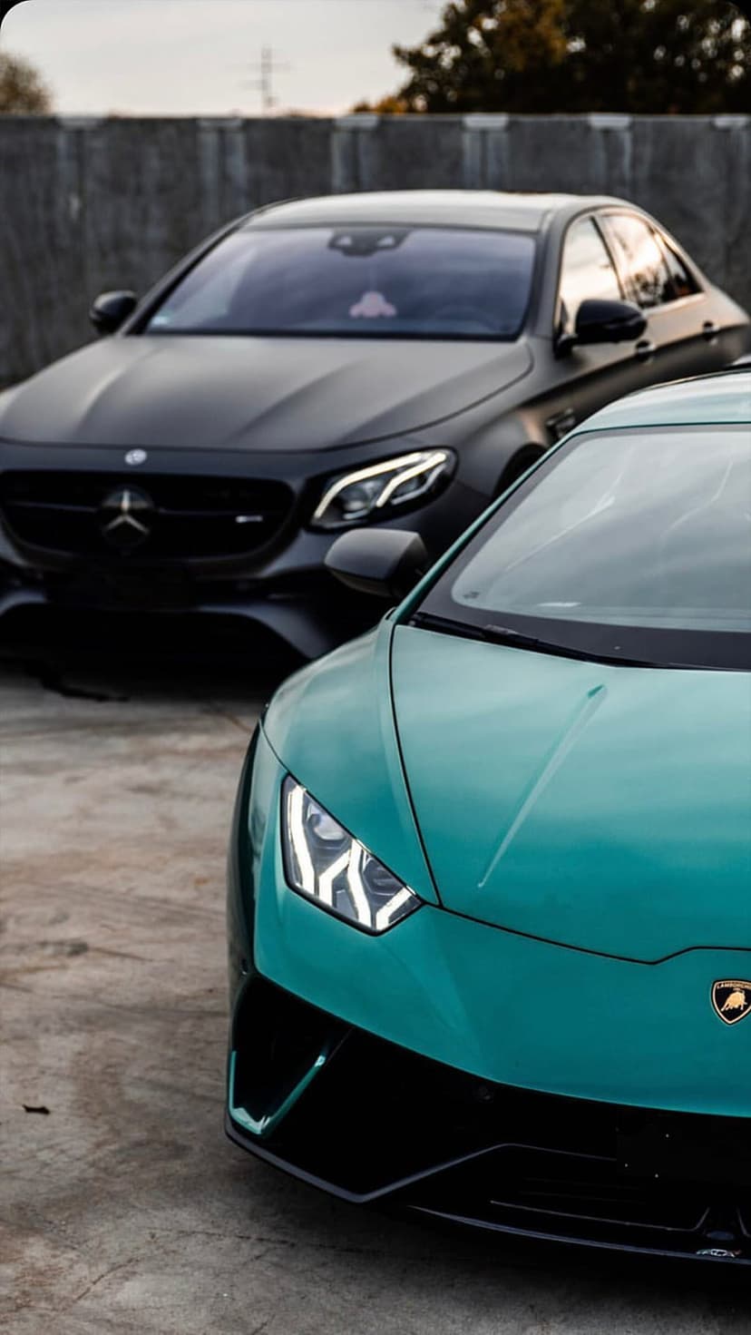 Teal Lamborghini Huracan and black Mercedes AMG sedan parked on a concrete surface against a textured wall.