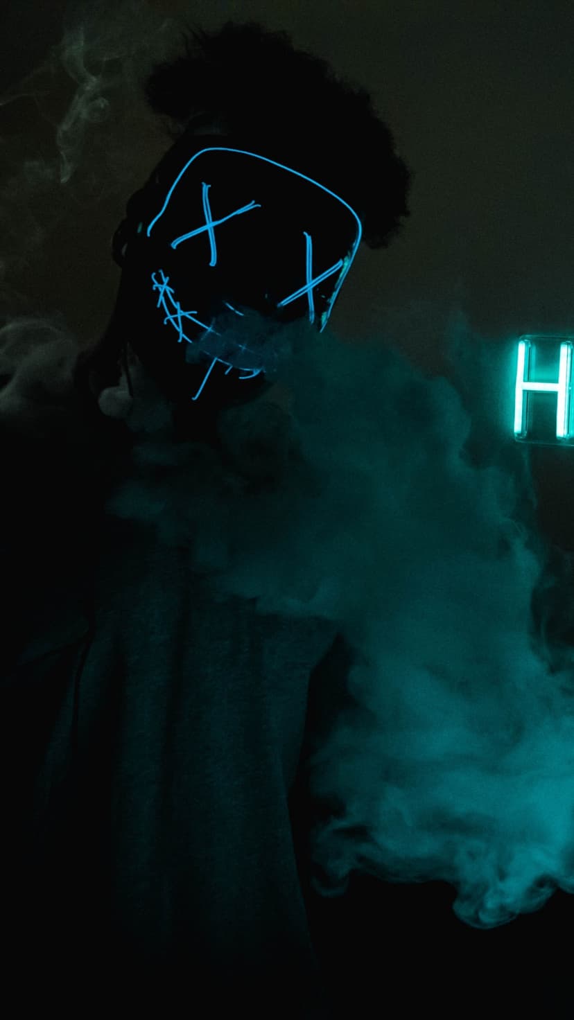 Masked figure with glowing blue X eyes and a stitched mouth stands within thick clouds of teal smoke at night.
